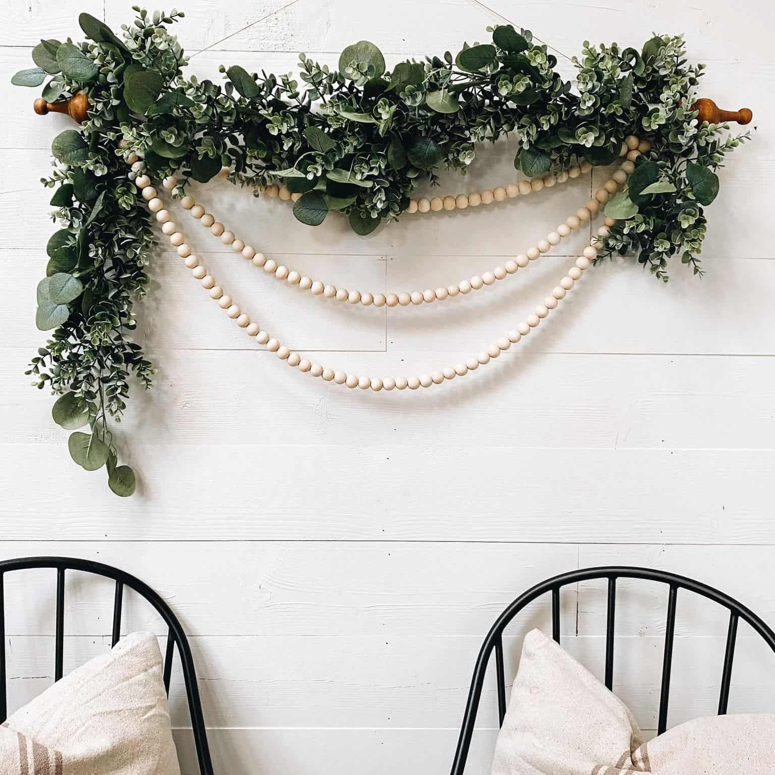Wooden rod draped with bushy leaves , a mix of eucalyptus and shrubbery with three strings of beige wooden beads hanging from end to end of the rod in different lengths. 